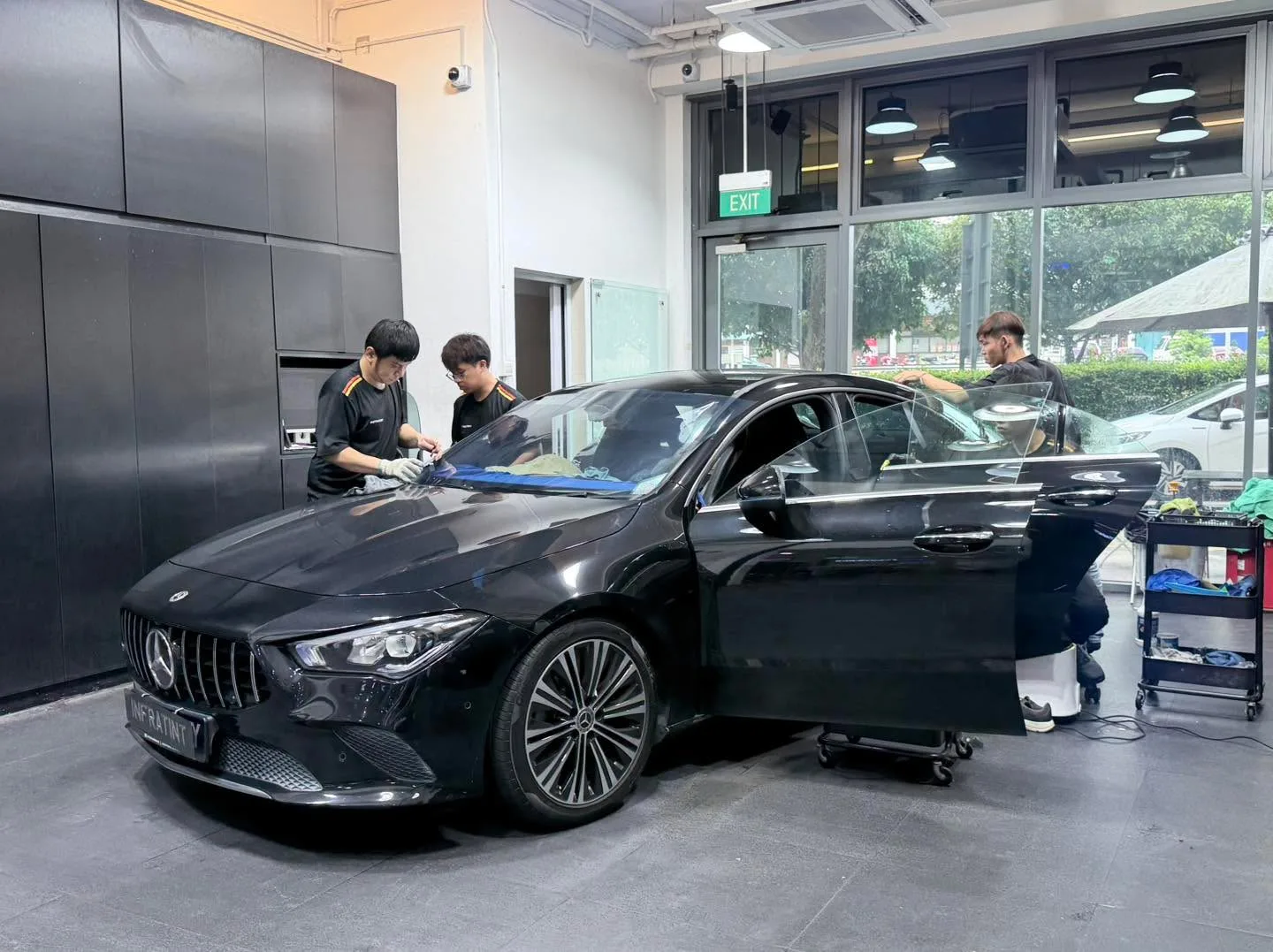A black Mercedes-Benz sedan with doors open, technicians applying window film to multiple windows.