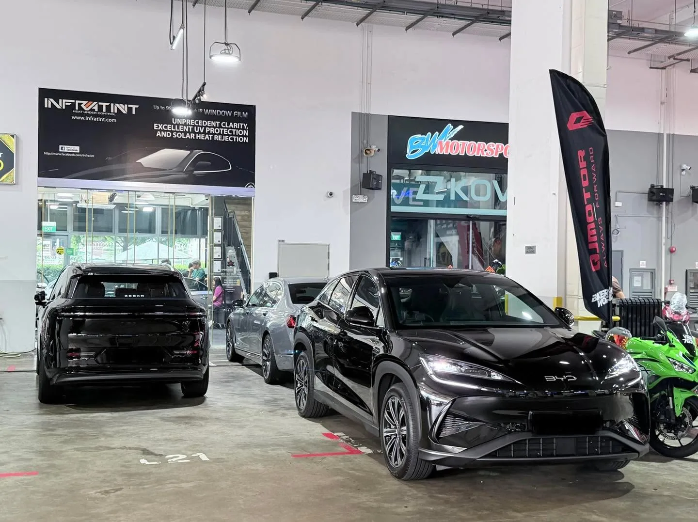 A black SUV, a silver sedan and a black electric vehicle parked in the garage with a green motorcycle visible to the right.