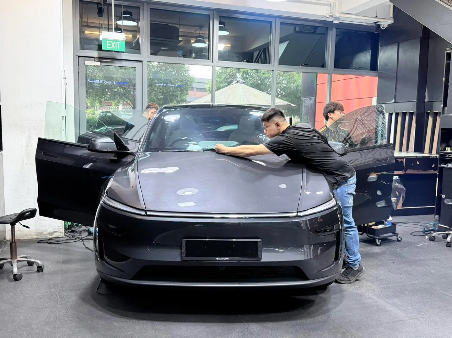 A grey sedan with doors open, technician applying film to the windscreen.