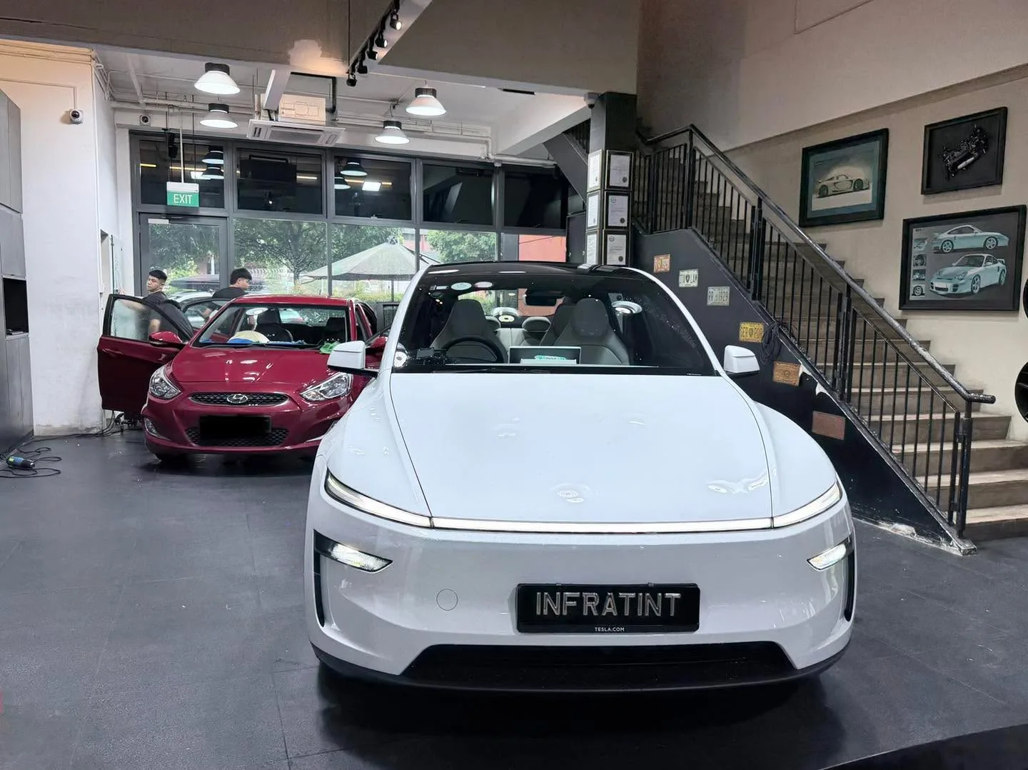 A silver Tesla sedan with open doors inside the workshop, with a maroon Honda sedan visible to the left.