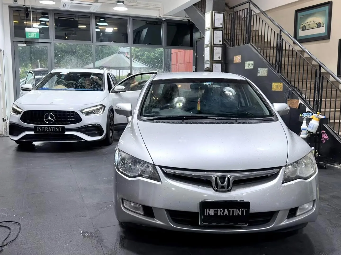 A silver Honda sedan in the foreground with a white Mercedes sedan behind it inside the workshop.