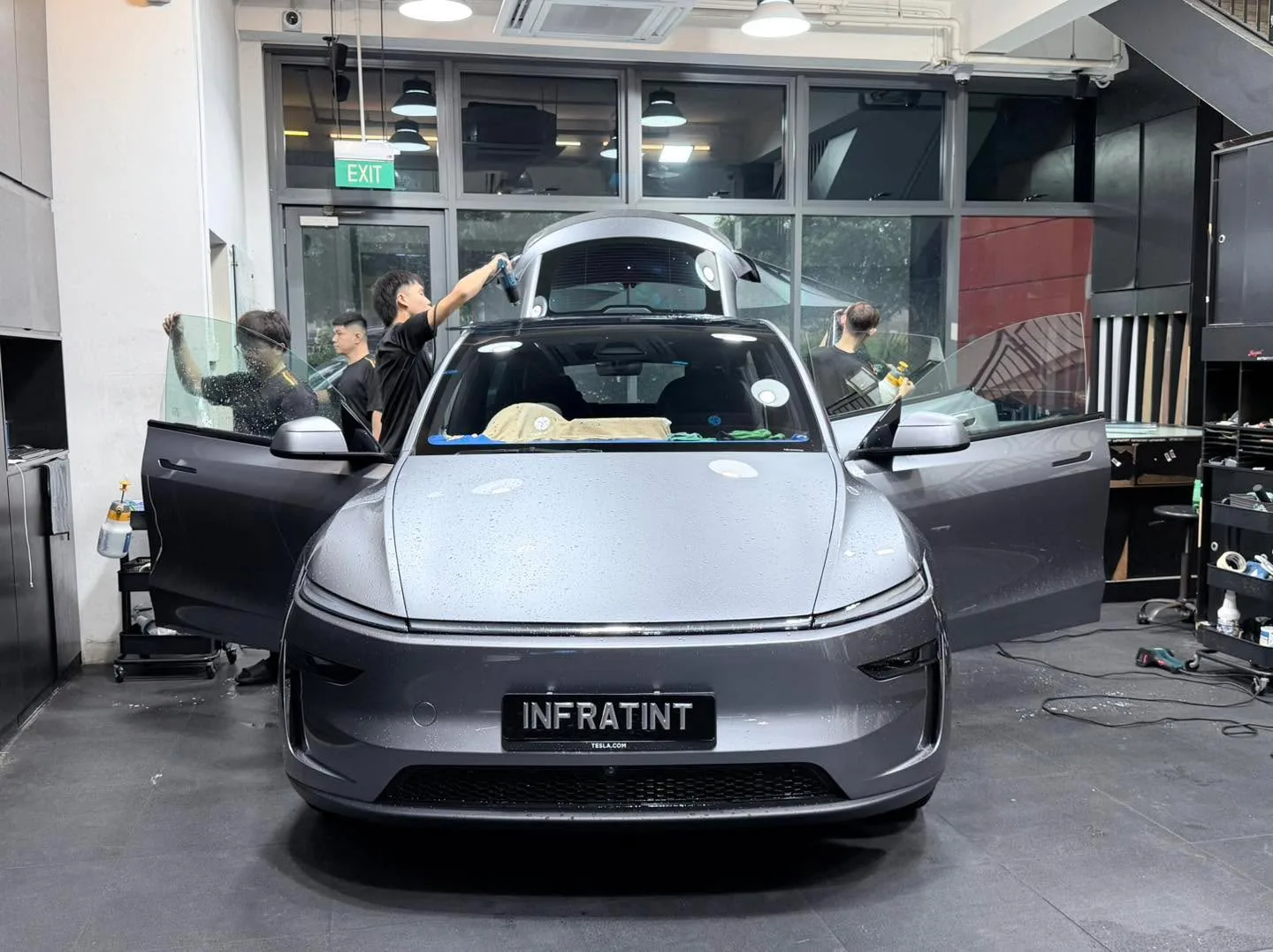 A silver Tesla sedan with open doors and technicians applying window tint, viewed from the front.