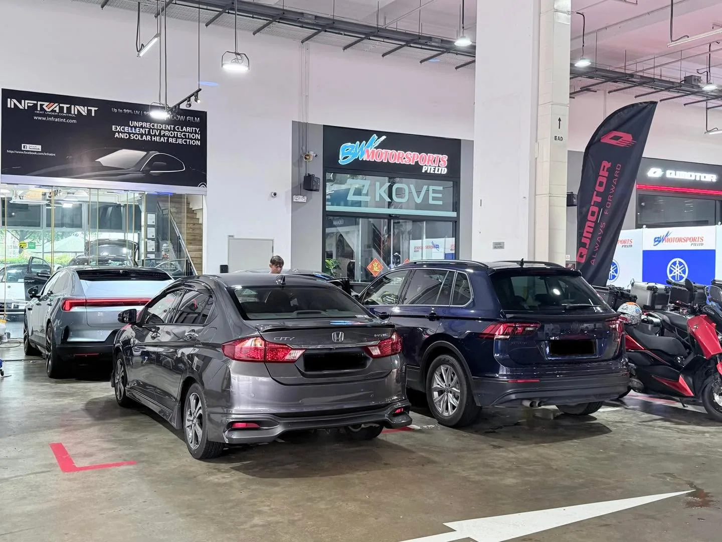 Three sedans and an SUV parked in the workshop bay. A grey sedan is in the foreground, a dark blue sedan in the centre, and a light-coloured SUV to the left.