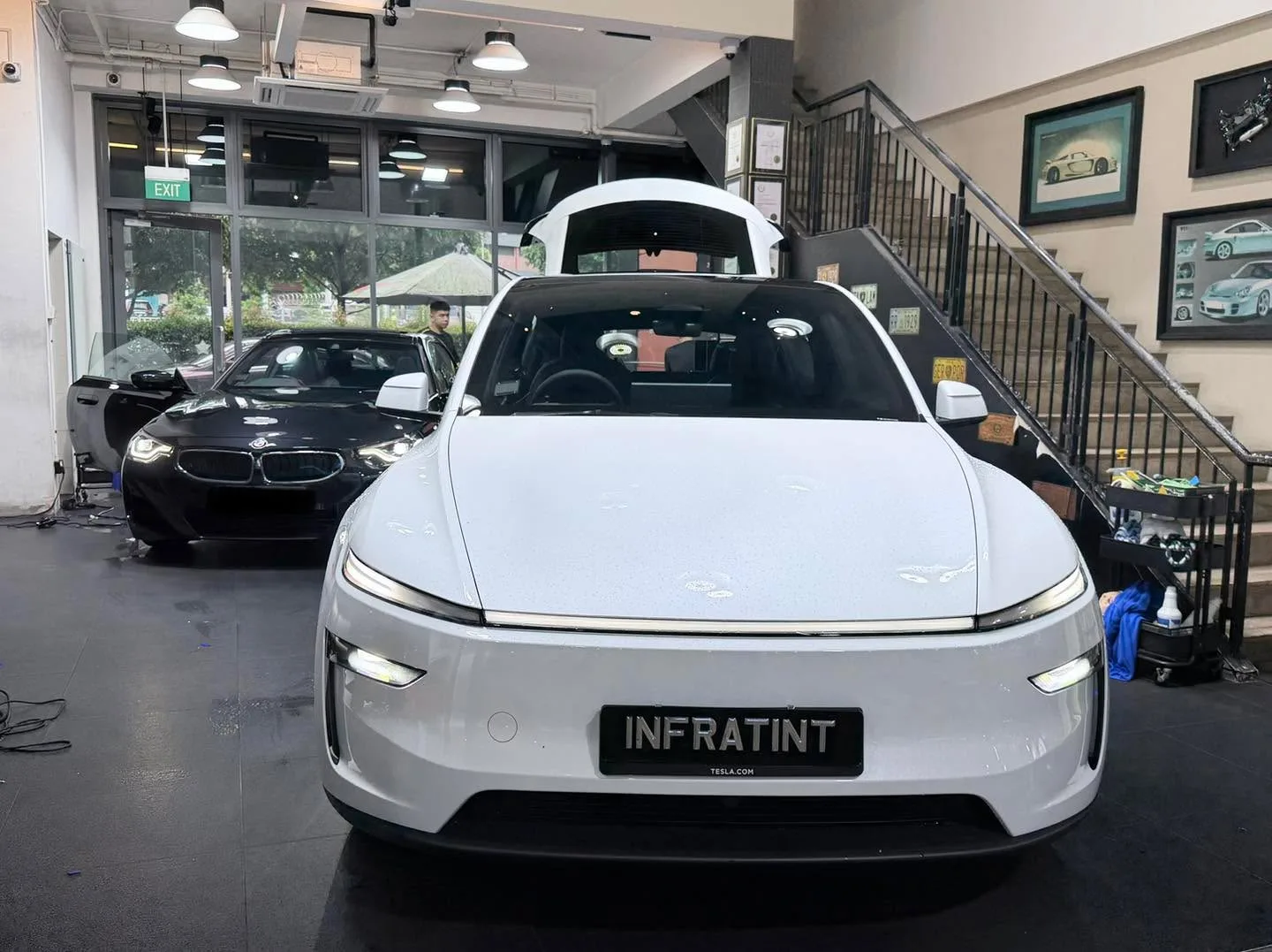 A white Tesla sedan displayed indoors with its front end facing the camera, parked in front of a black BMW sedan.