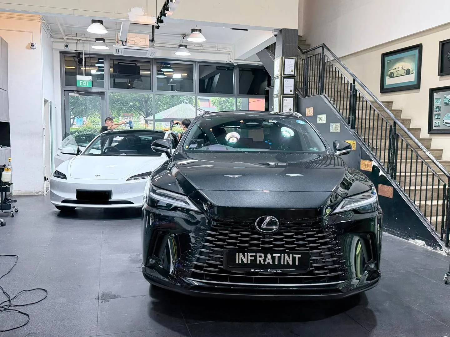 A black Lexus SUV with distinctive grille design in the foreground, with a white Tesla sedan visible behind it indoors.