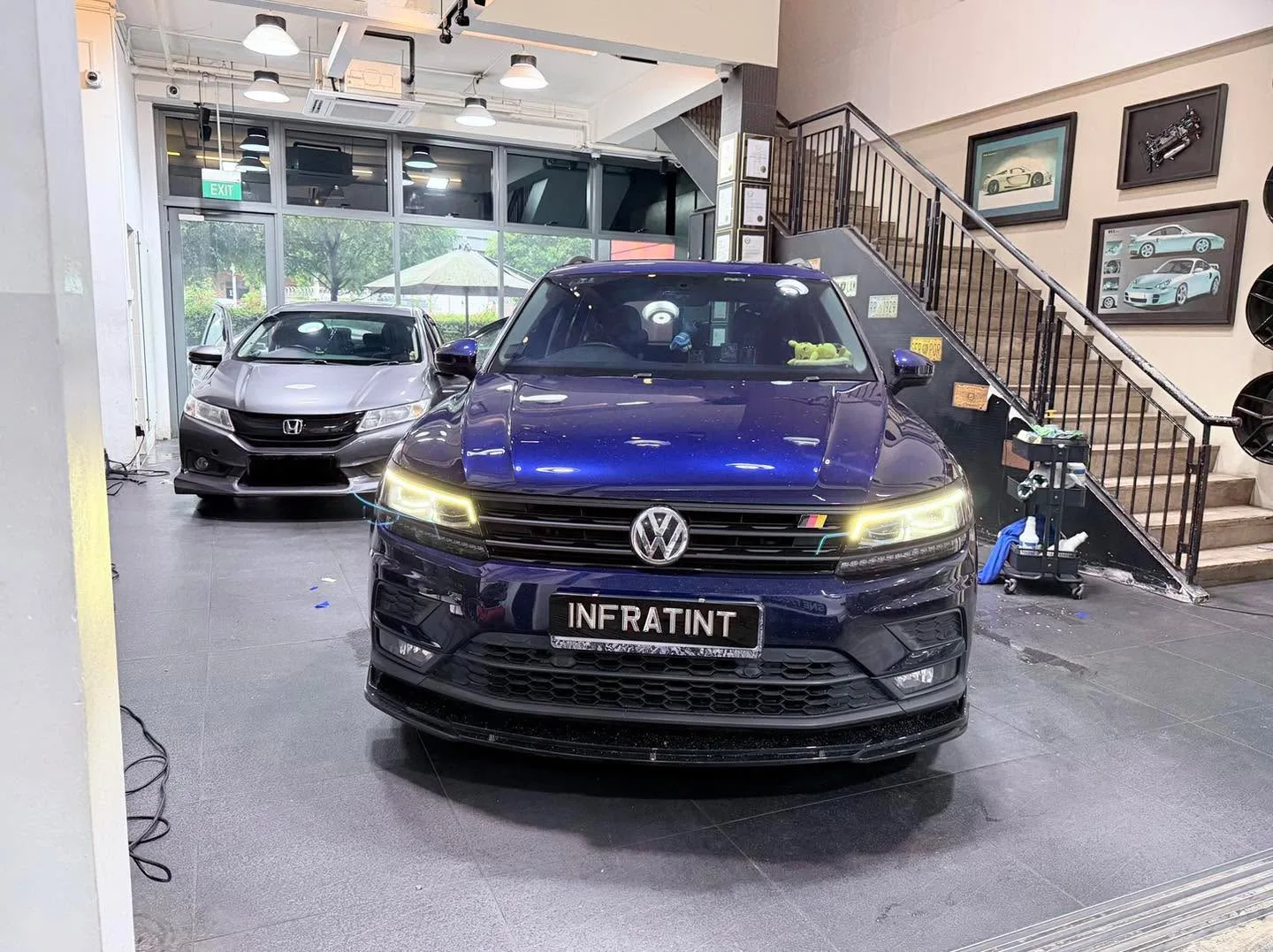 A dark blue Volkswagen SUV in the foreground with LED headlights illuminated, and a grey Honda sedan to the left, both indoors.
