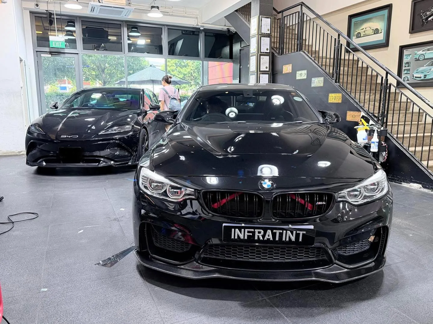 A black BMW M4 with prominent kidney grille facing the camera, and a black Tesla behind it.
