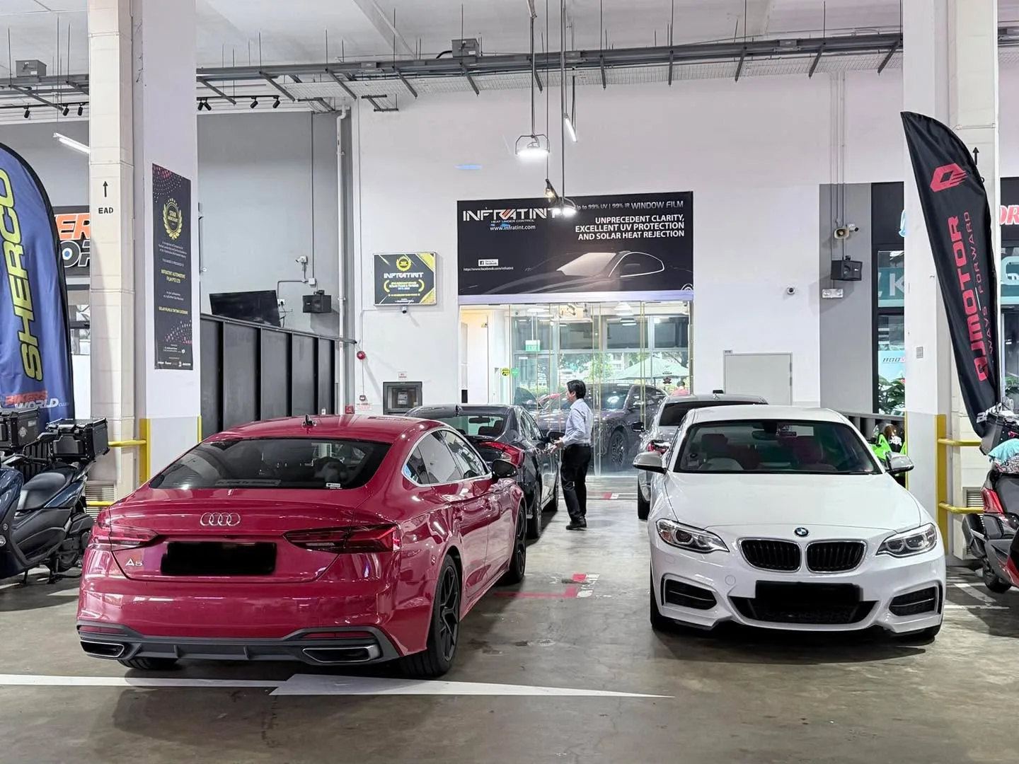 A red Audi sedan and a white BMW coupe parked side by side in a large workshop.