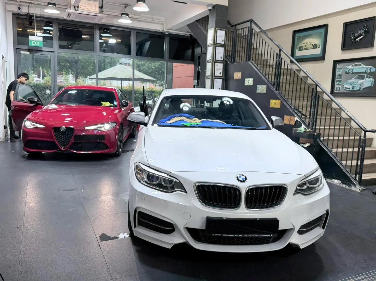 A red Alfa Romeo and white BMW coupe parked in a workshop with staff nearby.