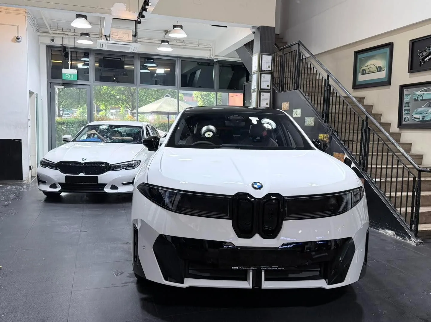 Two white BMWs parked indoors. The front car shows the distinctive grille and M Sport styling.