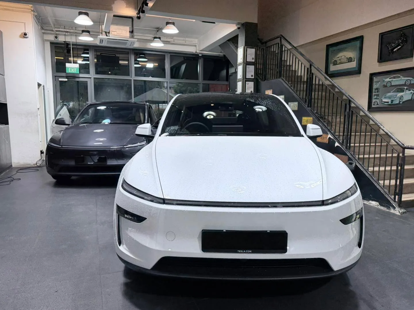 A white Tesla sedan viewed from behind, with a dark vehicle beside it.