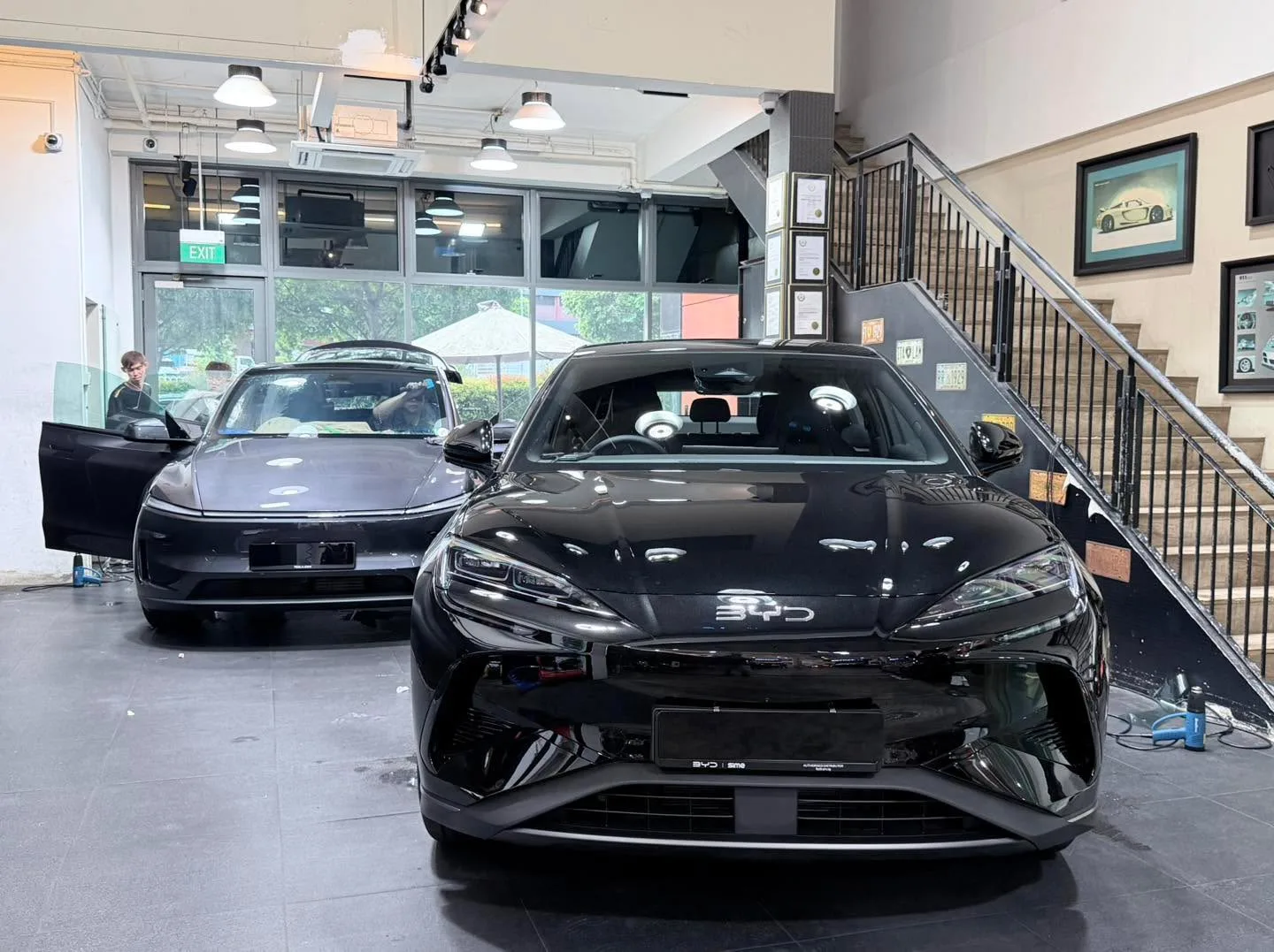 A black Tesla sedan and a white Tesla sedan parked indoors with technicians present.