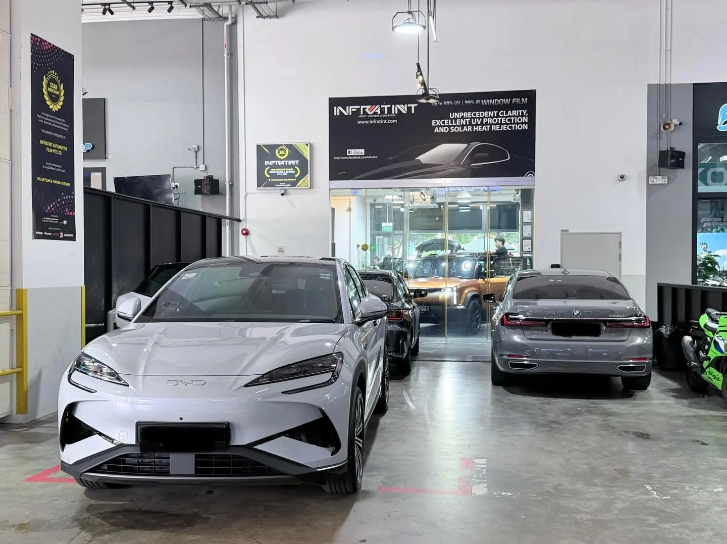 A white Jaecoo J6 SUV in the foreground and a grey-coloured saloon to its left in the workshop.