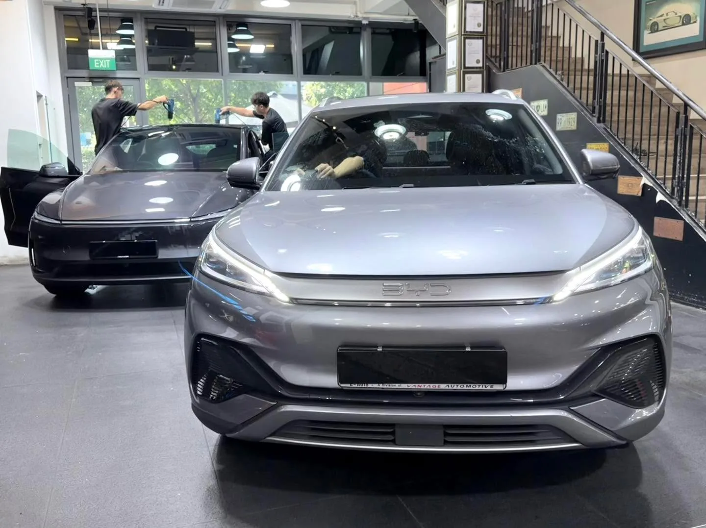 A grey BYD Sealion 7 saloon in the foreground with a black Tesla to its left, both indoors.