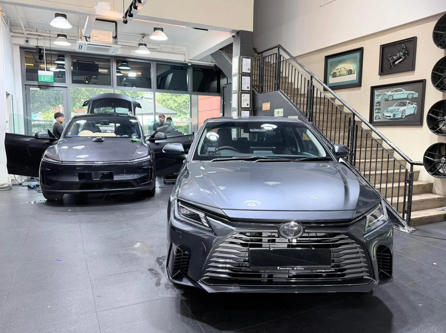 A silver Toyota Vios compact sedan and a black Tesla hatchback in the workshop with technicians nearby.