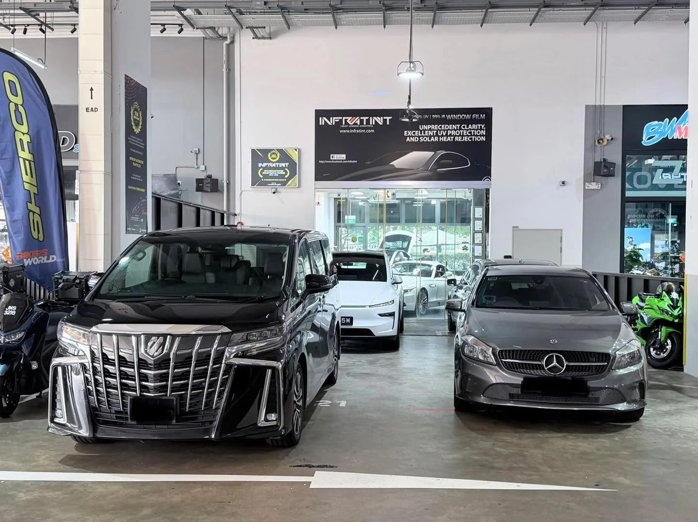 A black Toyota Alphard, a white Tesla, and a grey Mercedes-Benz parked in the workshop showroom area.