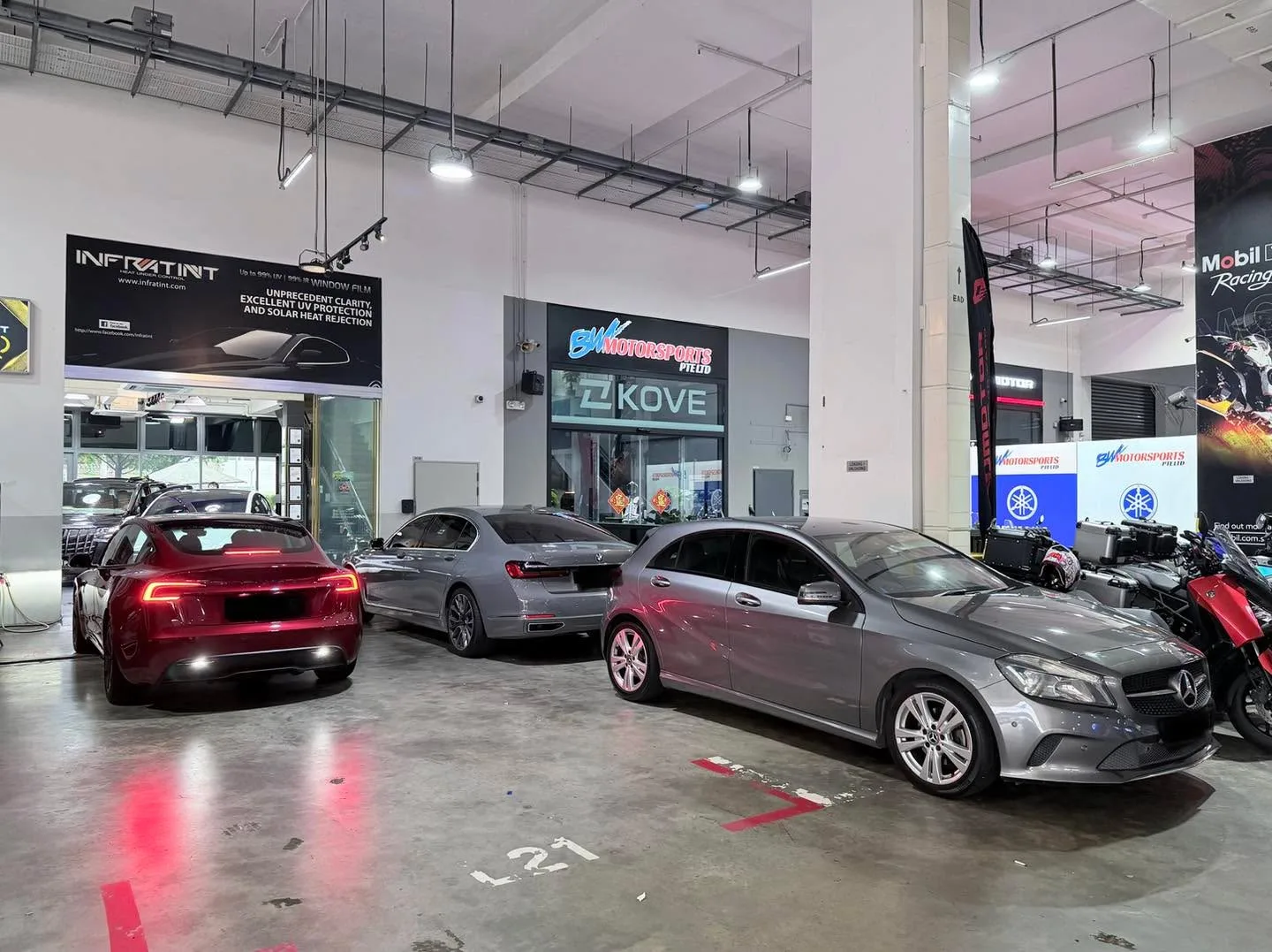 A red Tesla, a grey sedan, and a dark red Mercedes-Benz at the workshop in the evening.