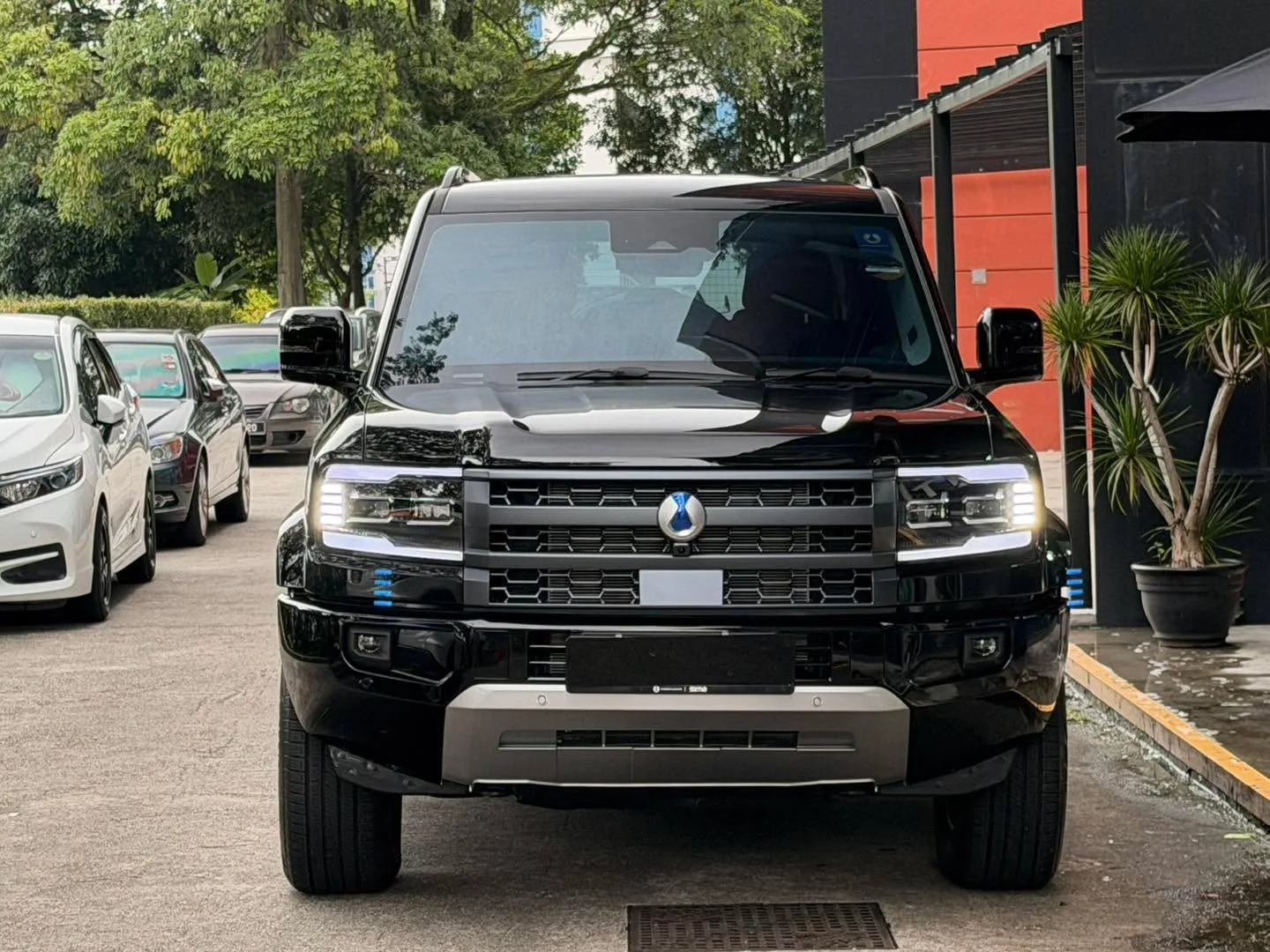 A black Denza B5 SUV parked at the workshop, viewed from the front, with other vehicles and greenery in the background.