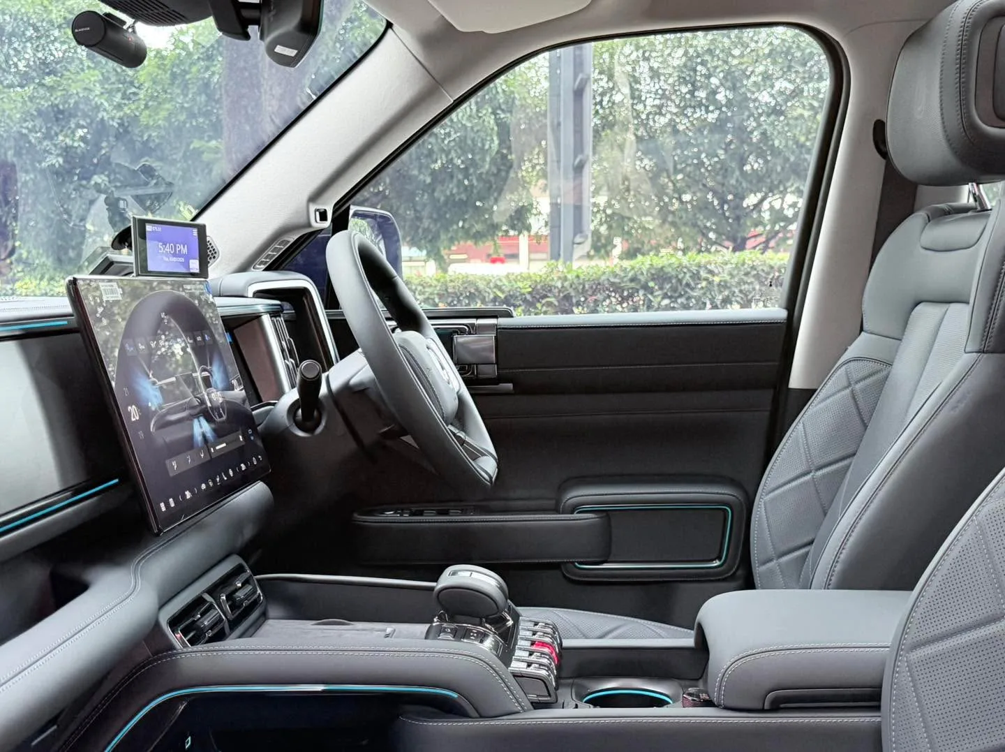 Interior of the Denza B5 showing the dashboard, steering wheel, digital display, and tinted side windows with a view of trees outside.