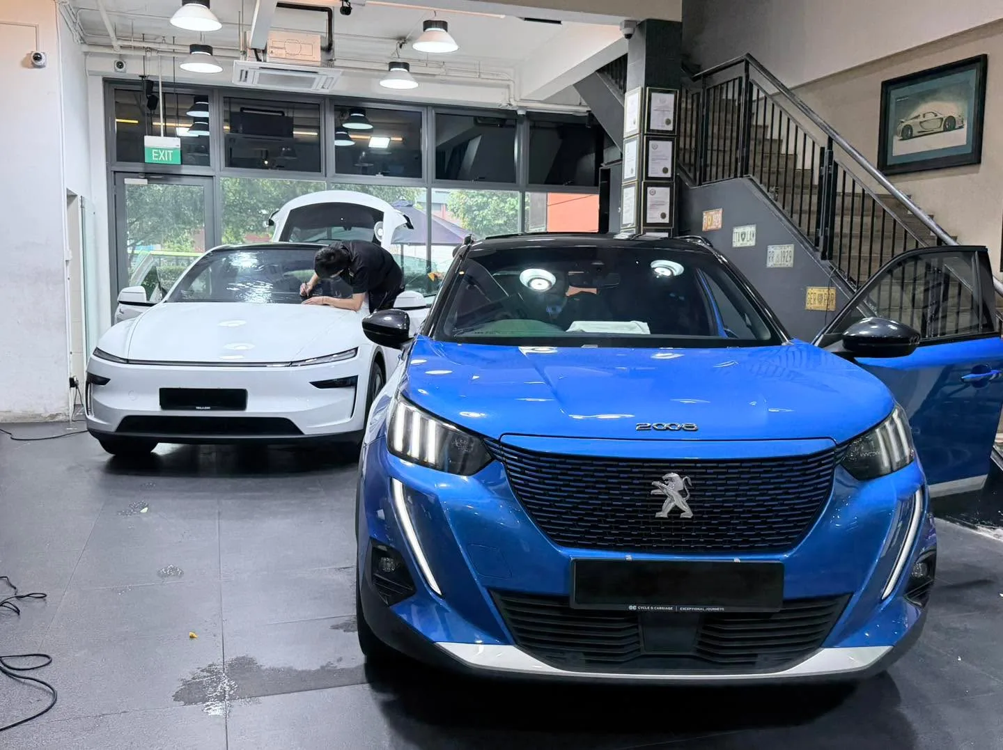 A blue Peugeot compact SUV and a white Tesla sedan at the workshop entrance.