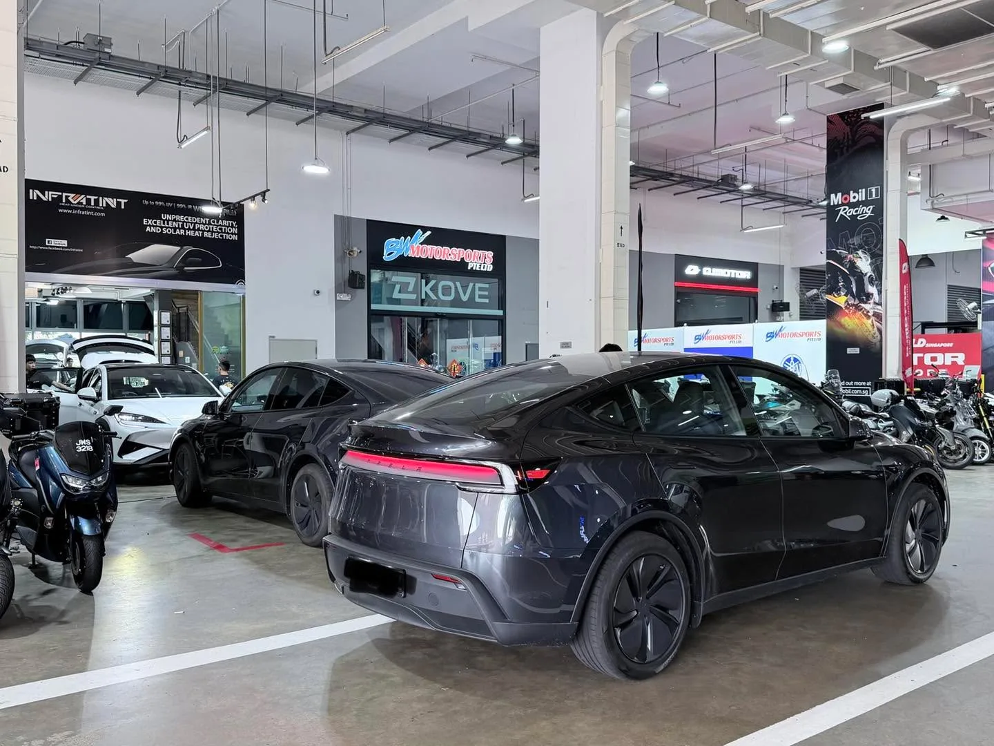 A dark-coloured Tesla Model Y and a white car parked in a workshop showroom.