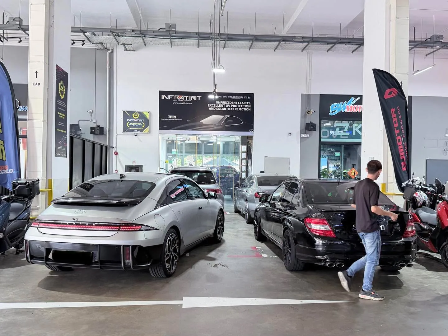 A white electric sedan and a black saloon inside the workshop with overhead lighting.