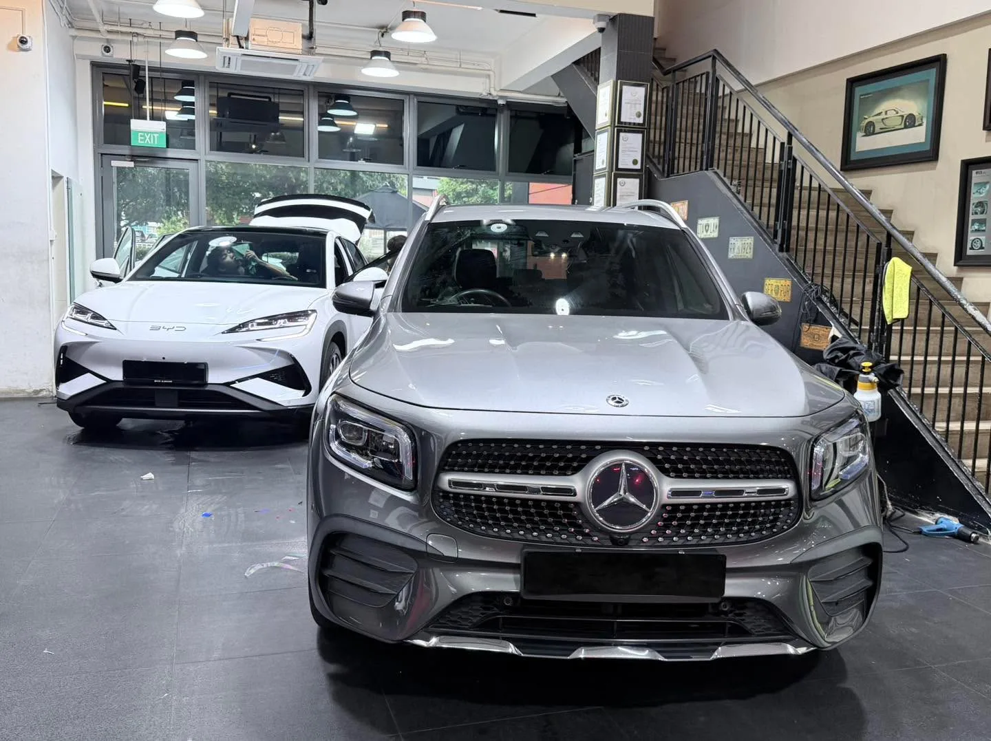 A white BYD Sealing 7 SUV and a silver Mercedes-Benz GLC SUV parked side by side indoors.