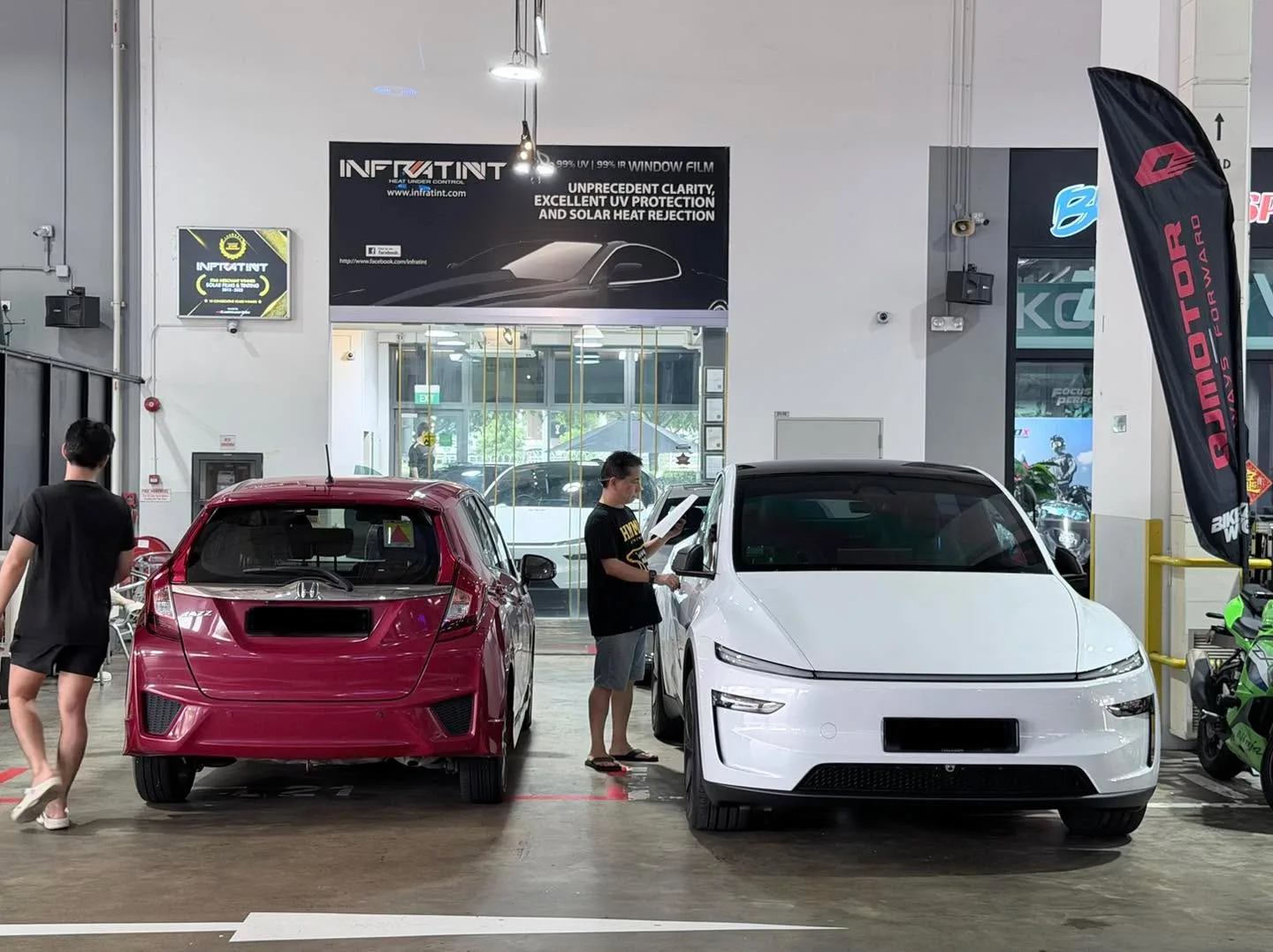 A red hatchback and white electric sedan undergoing window tinting indoors with technicians working nearby.