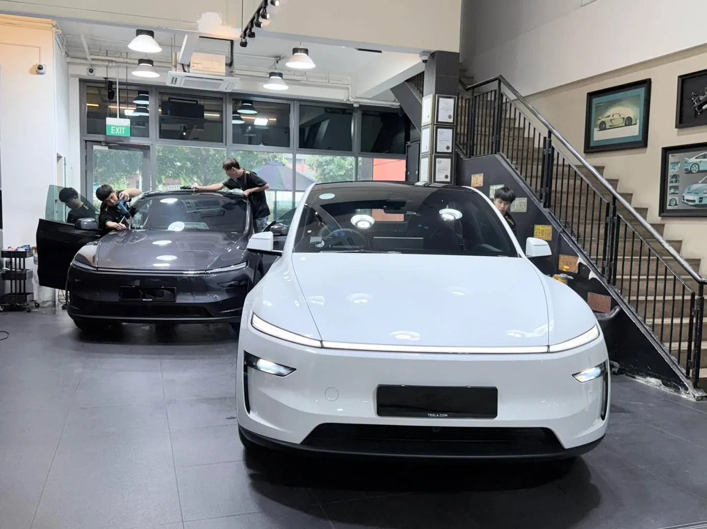 A white electric sedan in the foreground with tinted windows, a black electric sedan behind it being worked on by technicians.
