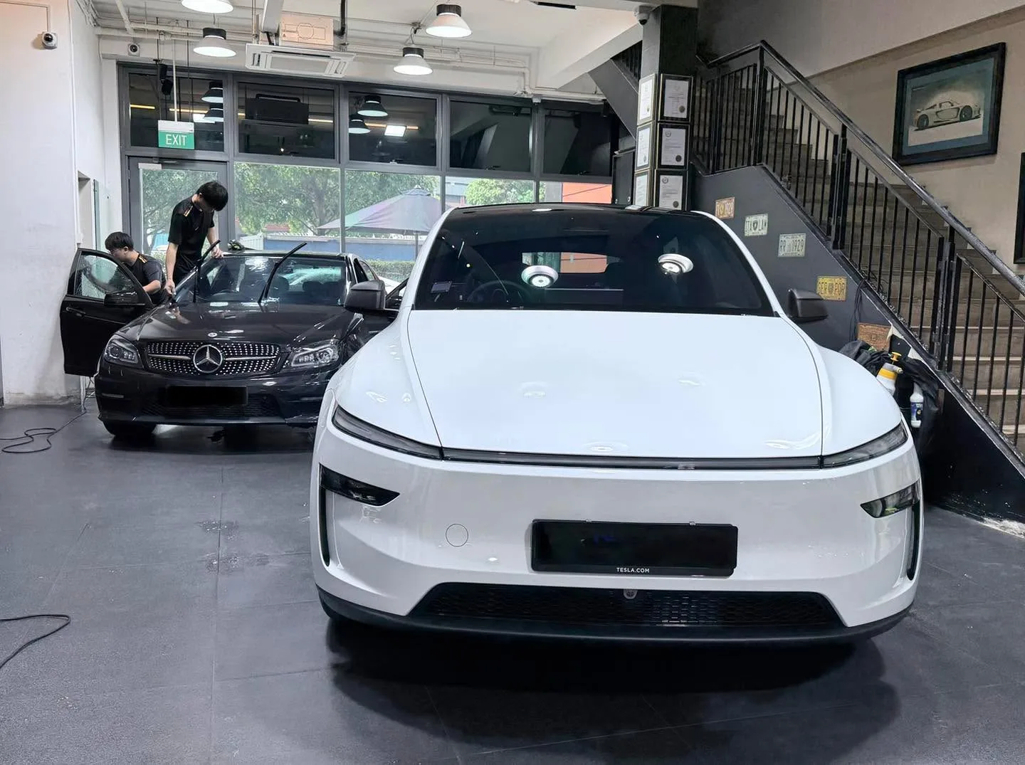 White electric sedan in foreground with black Mercedes-Benz to its left, both inside the workshop with technicians nearby.