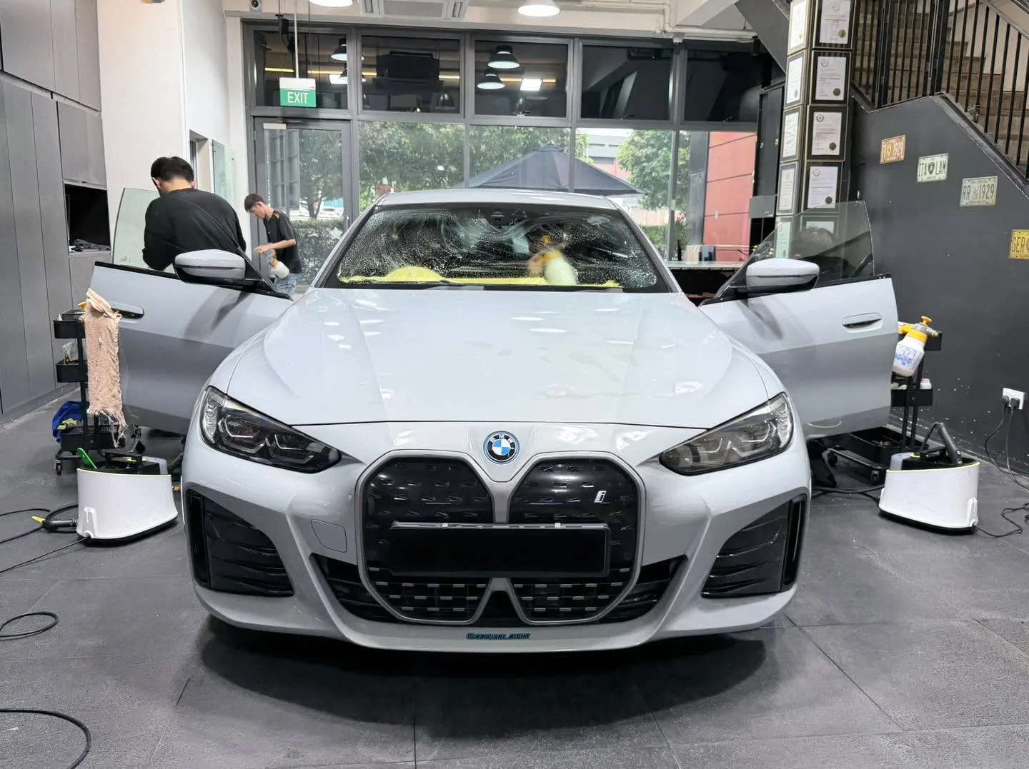 White BMW with distinctive kidney grille in foreground, technicians working on the vehicle, workshop interior visible.