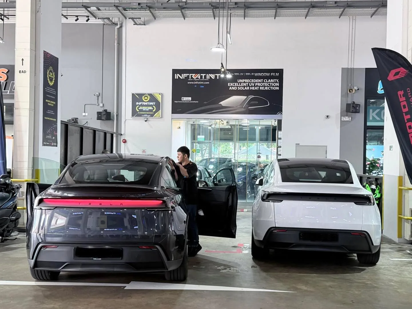 A black Tesla Model Y with distinctive red tail light bar and a white BYD Atto 3 crossover parked indoors at the workshop.