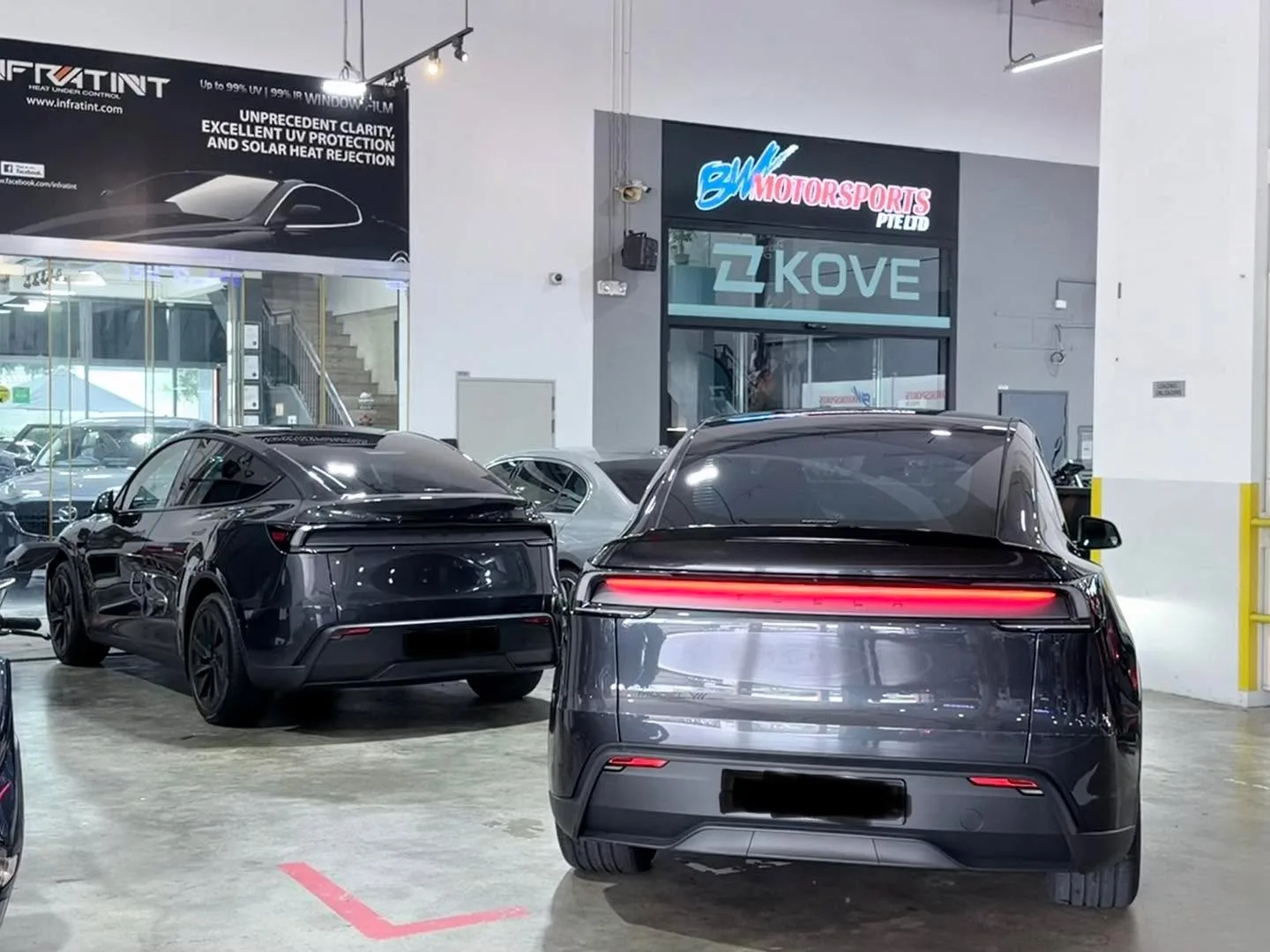 A black Tesla Model Y and another dark sedan parked in a workshop with yellow safety bollards.