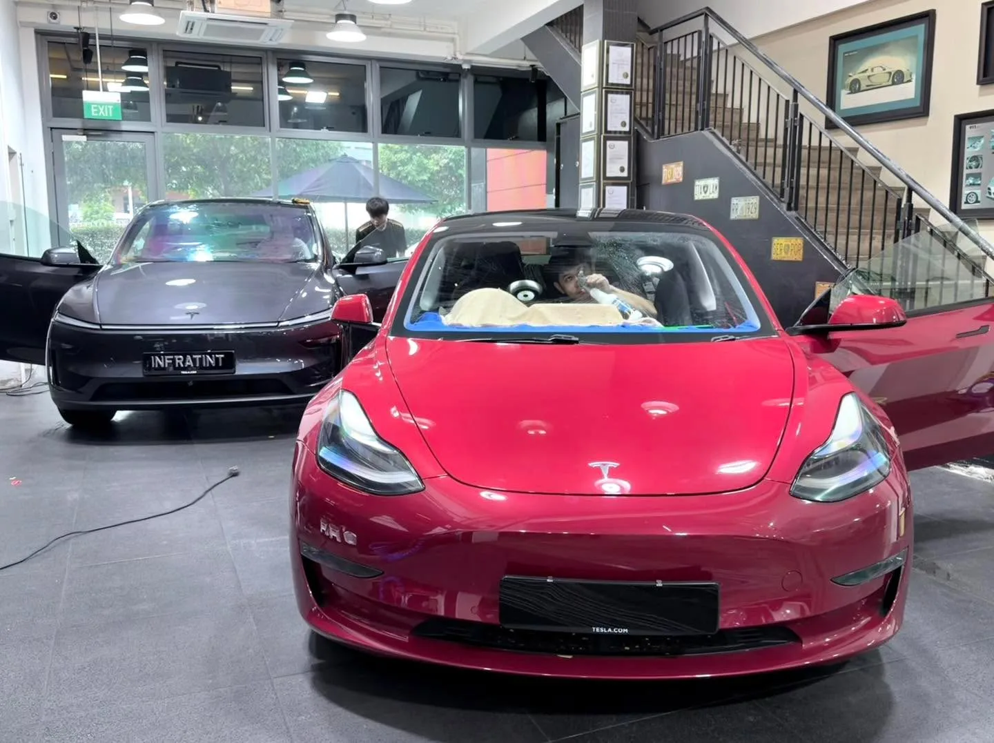 A bright red Tesla Model 3 and a dark Mercedes-Benz with open doors inside the workshop.
