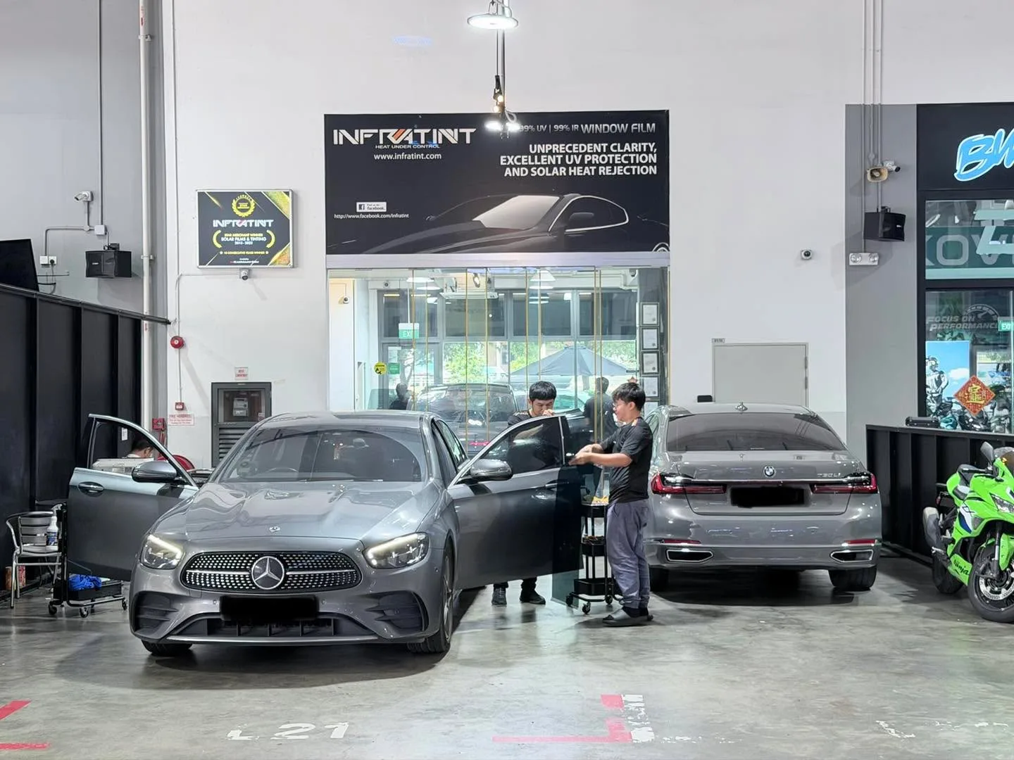 A grey Mercedes-Benz E-Class and a grey BMW 7 Series in the workshop, with technicians working nearby.