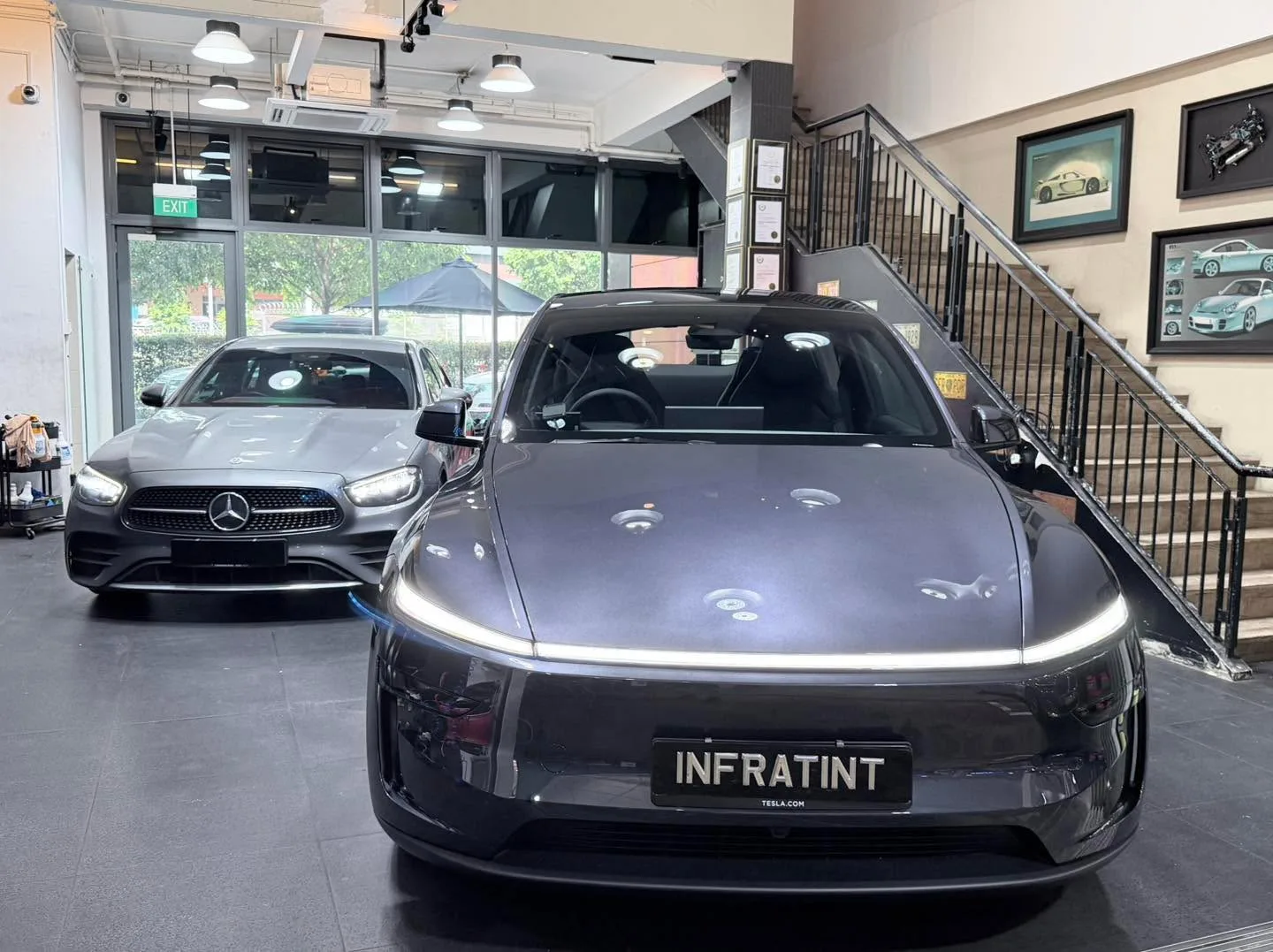 A dark grey Tesla sedan and a grey Mercedes-Benz E-Class parked in the indoor workshop.