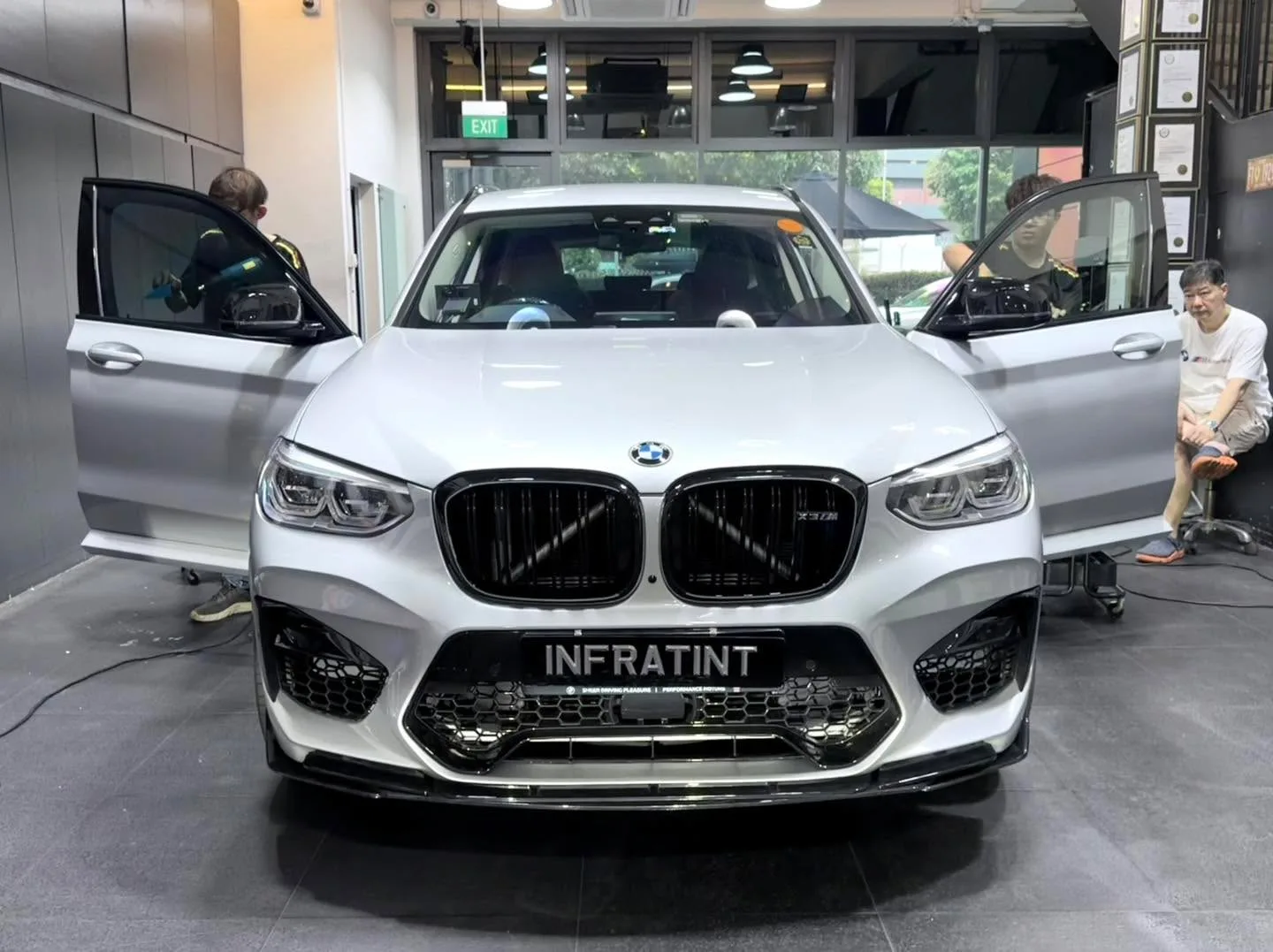 A white BMW X3 from the front with doors open in the workshop, technicians present.