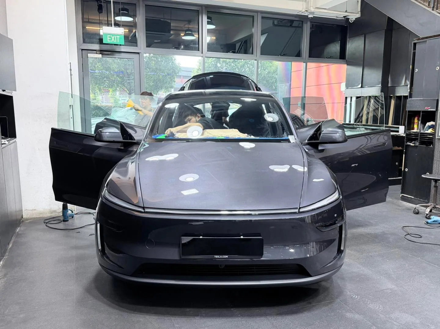 A dark grey Tesla hatchback from the front with doors open, technicians working inside the vehicle.