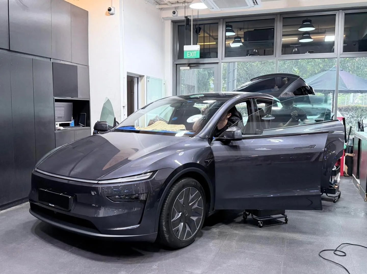 A dark grey Tesla sedan from the side with doors open, technician working on the window tinting.