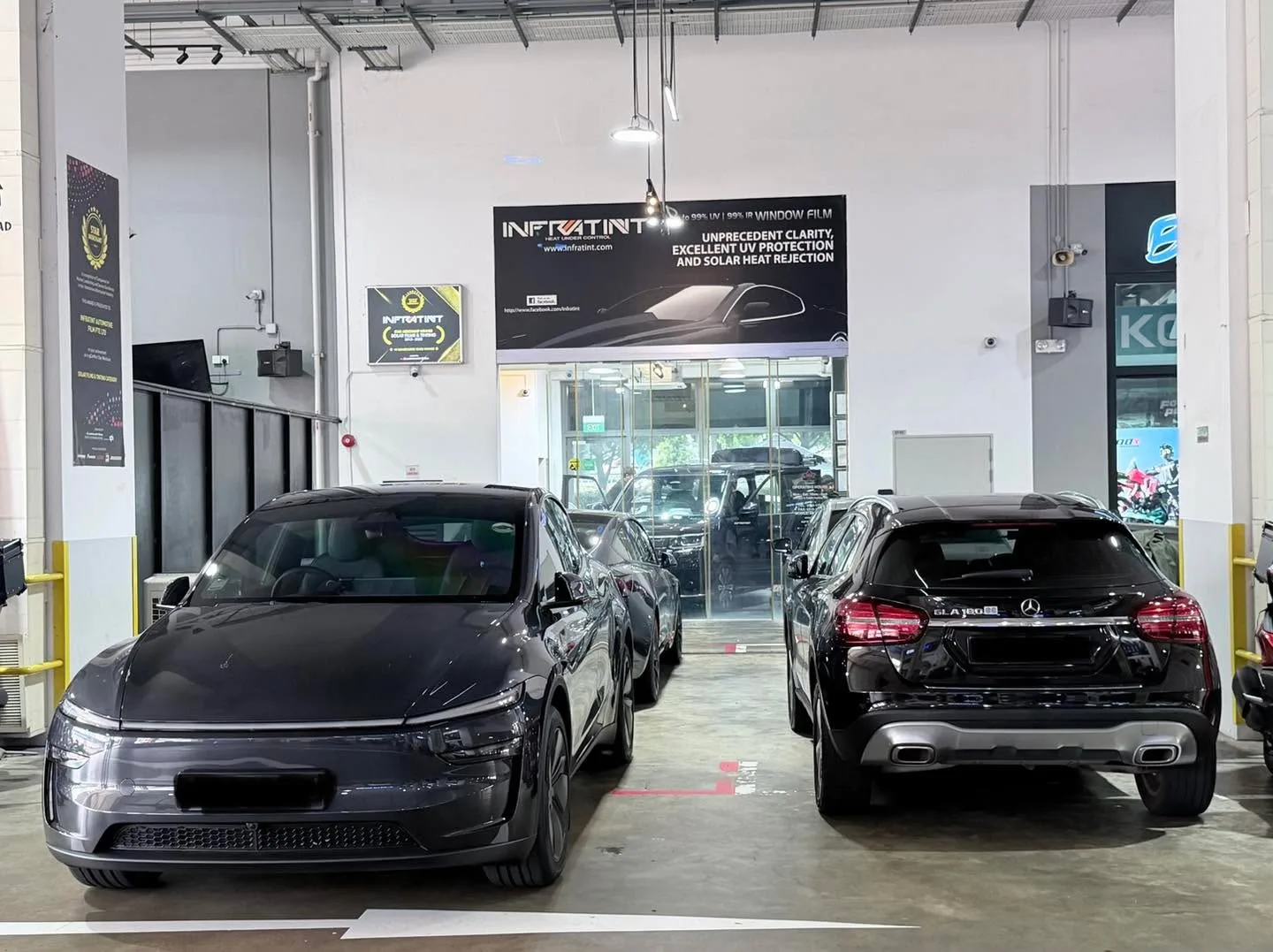 A grey Tesla sedan and a black Mercedes-Benz SUV parked indoors, with workshop frontage visible.