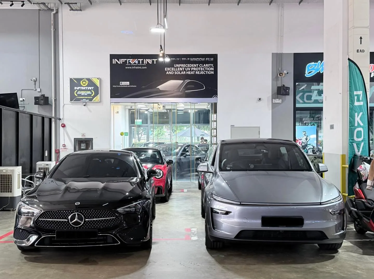 A black Mercedes-Benz sedan, a silver Tesla sedan, and workshop signage in the background.