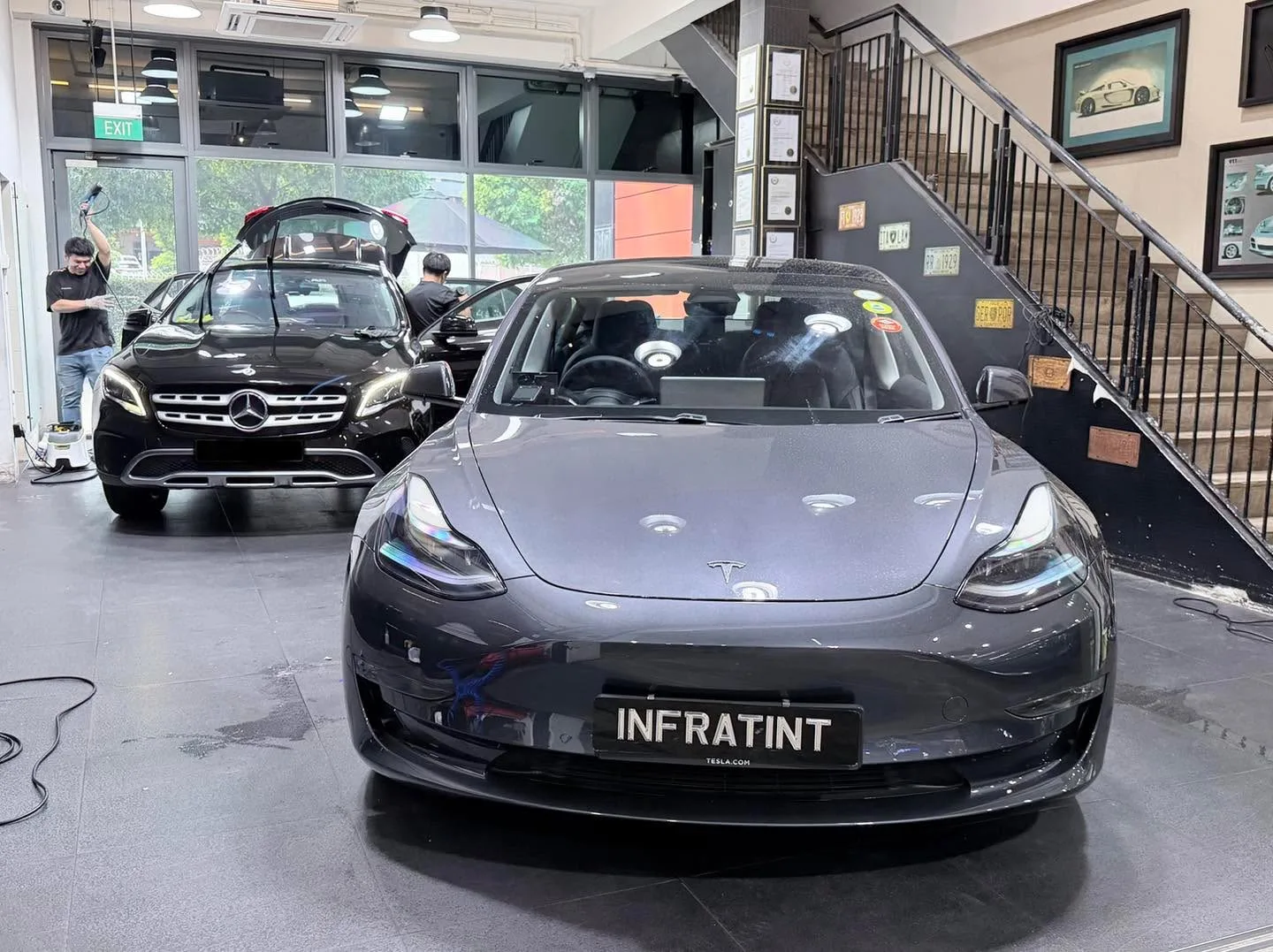 A dark grey Tesla sedan and a white Honda sedan parked indoors, with staff working nearby.