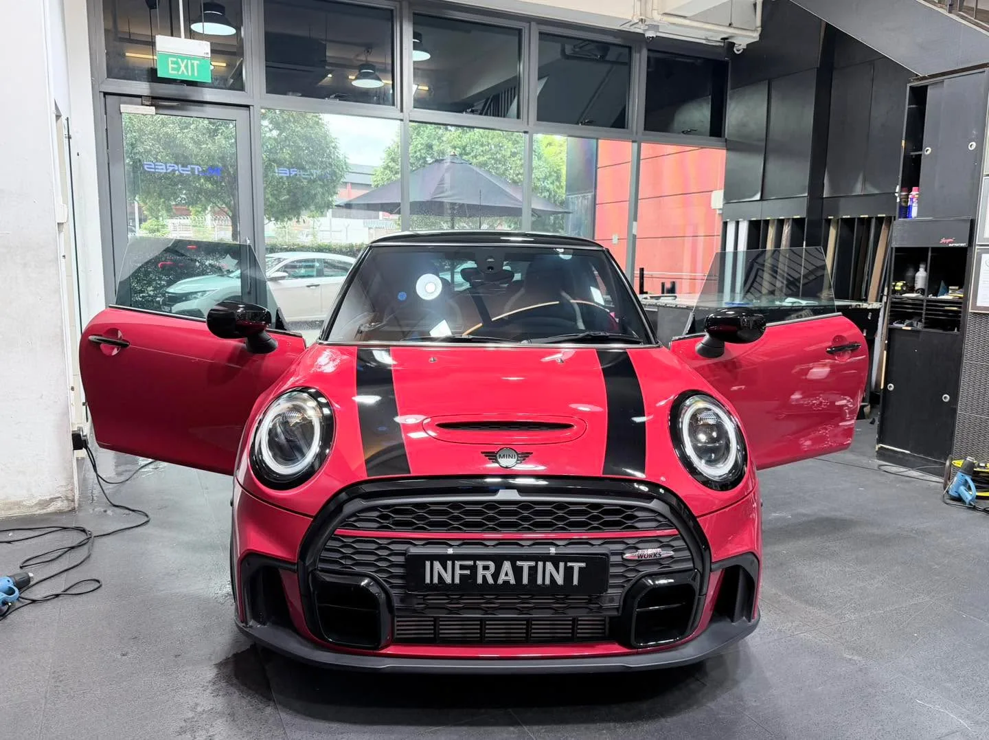 A red Mini hatchback with black stripes and open doors, parked indoors with shop windows visible.