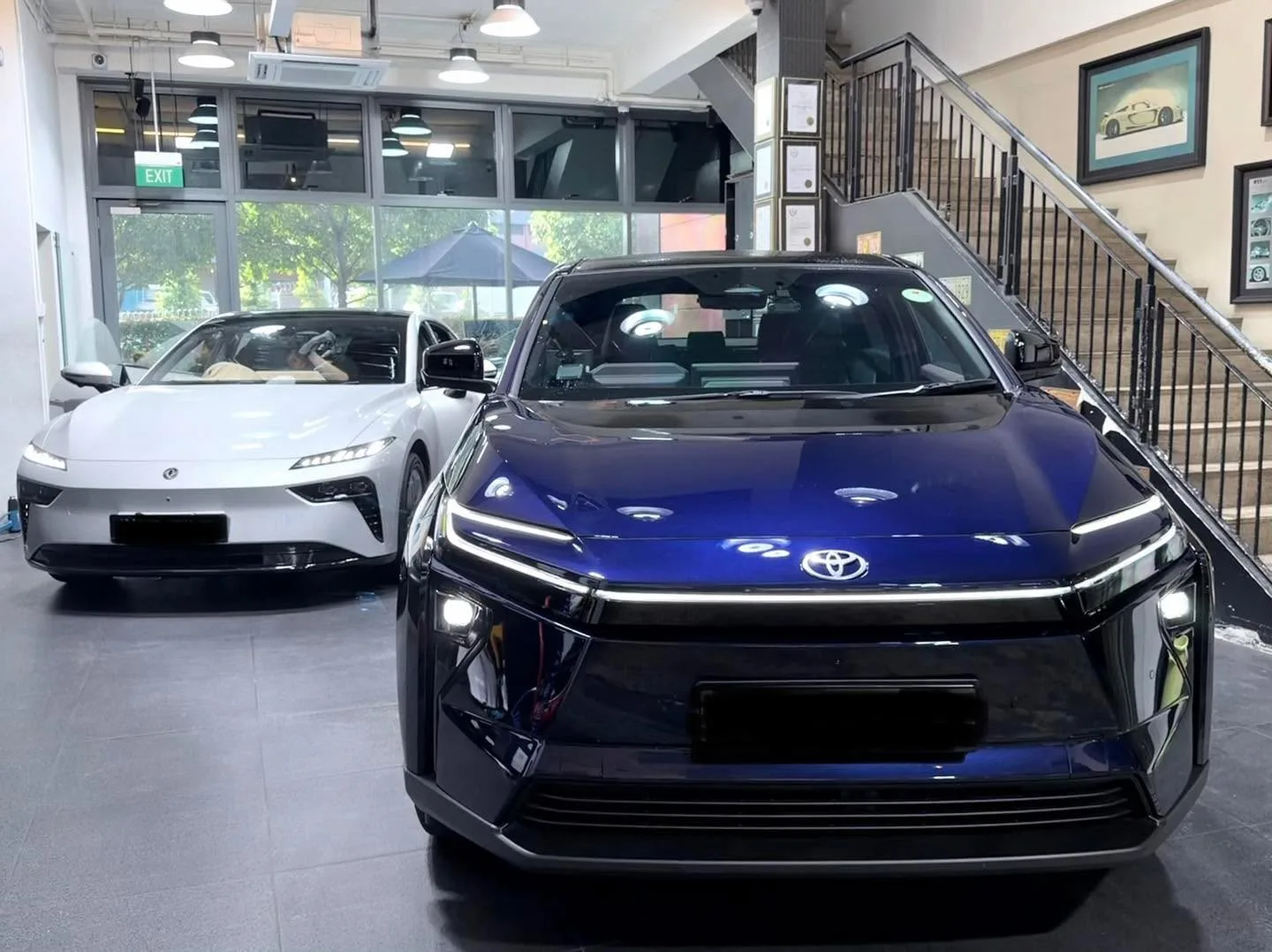 A dark blue electric saloon in the foreground and a white Tesla saloon beside it inside the workshop.