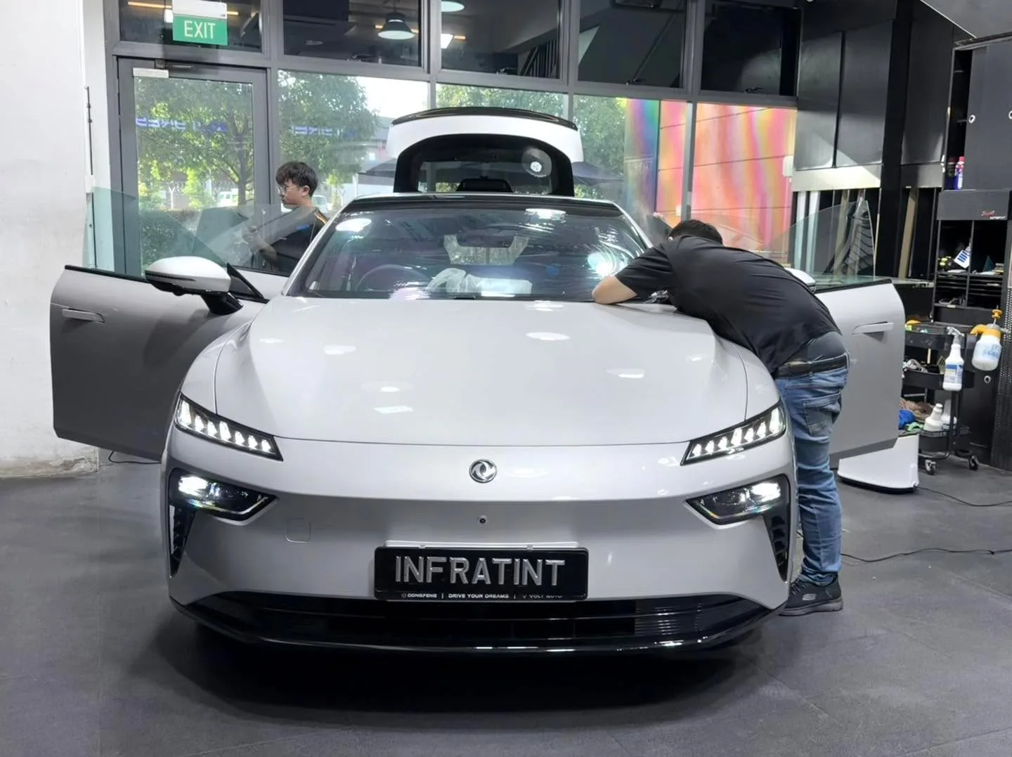 A white Dongfeng 007 sedan inside the workshop with its driver's door open, technician working on window tinting.