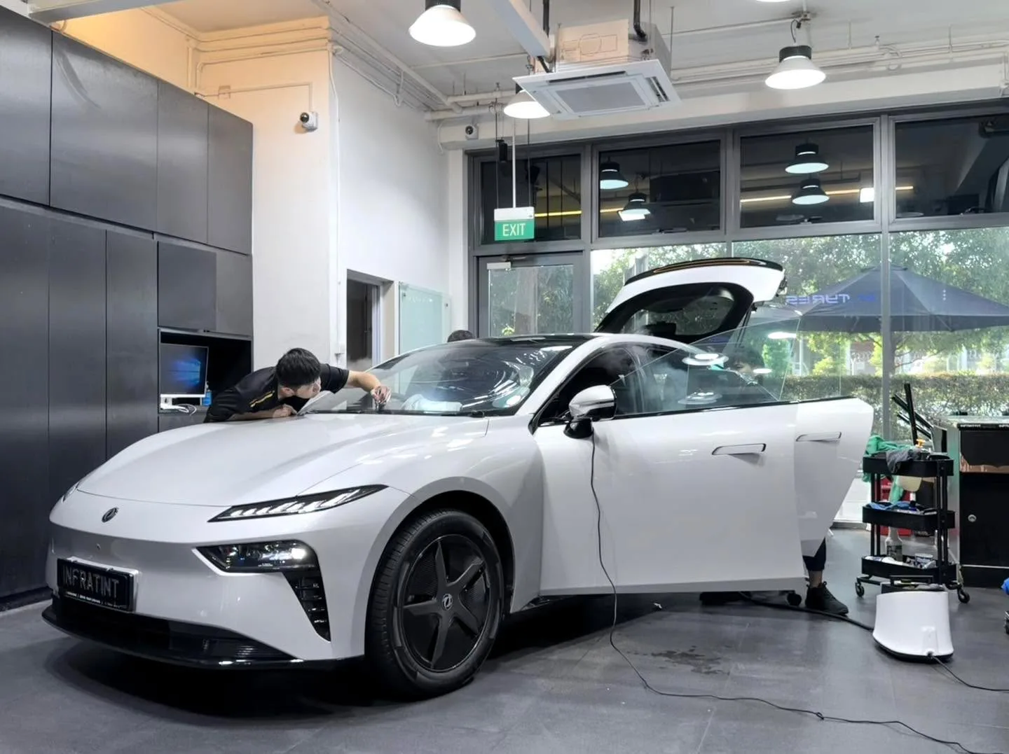 A white Dongfeng 007 sedan inside the workshop with both front doors open, technician applying tint to the windscreen.