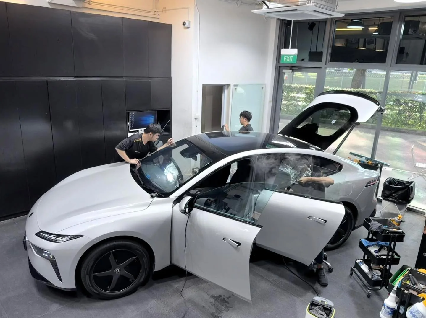 A white Dongfeng 007 sedan inside the workshop with doors open, showing tinting work in progress on the side windows.