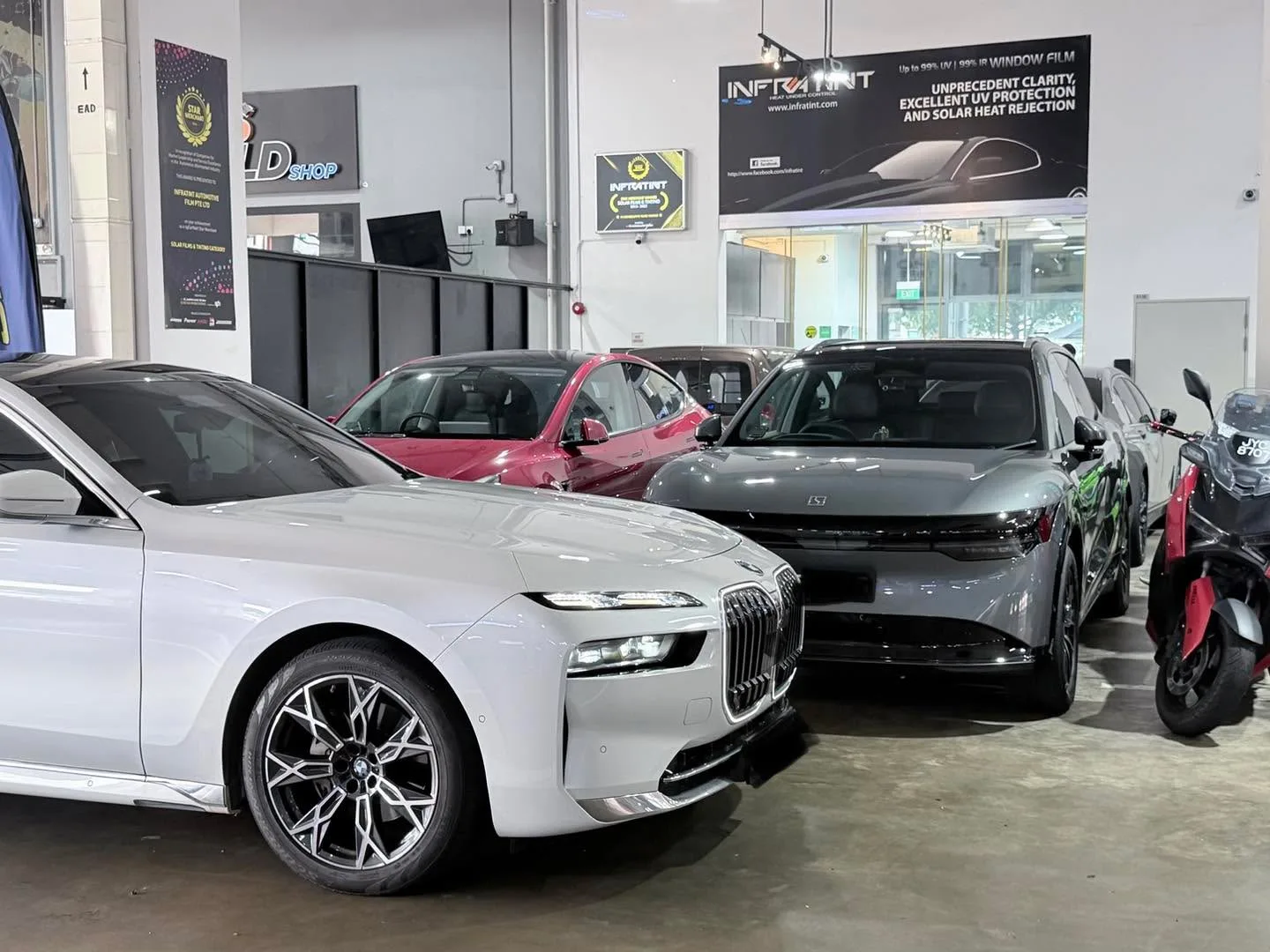 White BMW 7 Series sedan with distinctive kidney grille, silver Fiat hatchback, red Tesla Model Y, and grey sedan in workshop.
