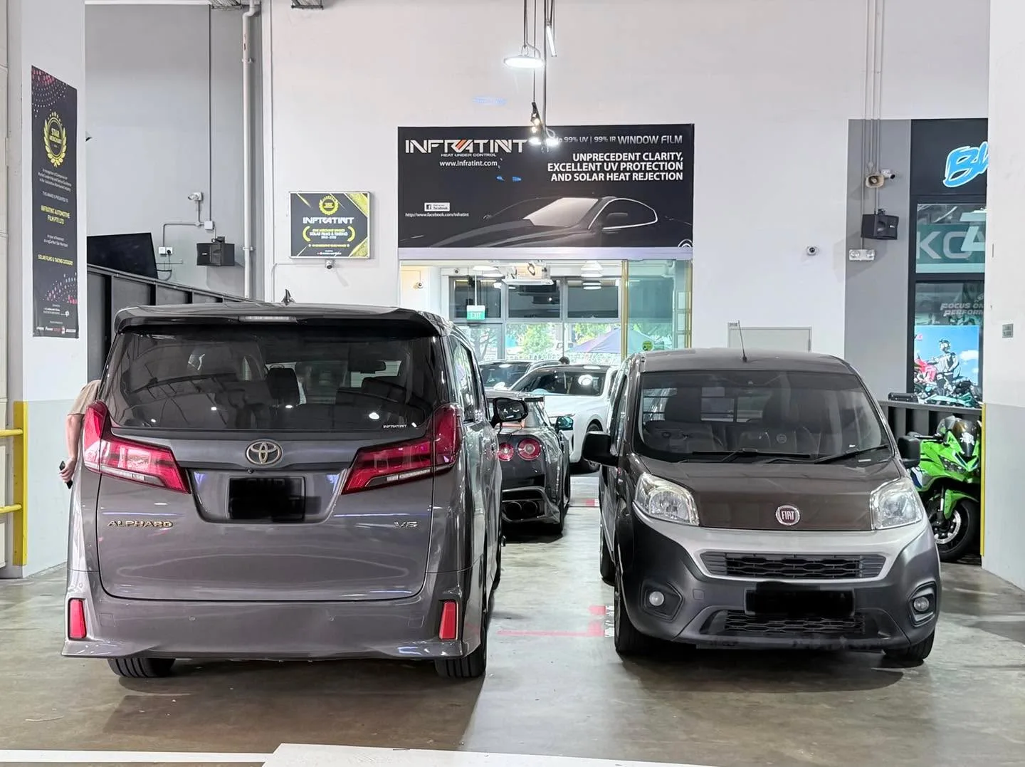 Rear three-quarter view of grey Fiat Forino hatchback beside a brown Toyota Alphard minivan in workshop.