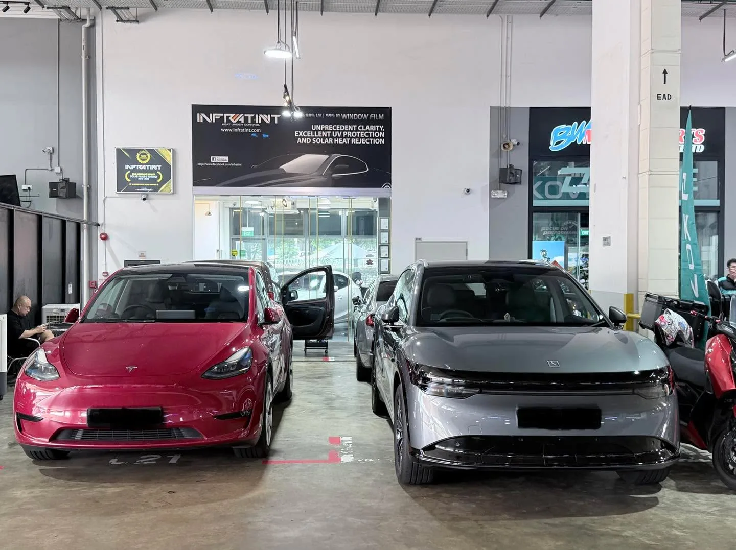 Red Tesla Model Y sedan next to grey BMW saloon in workshop space.